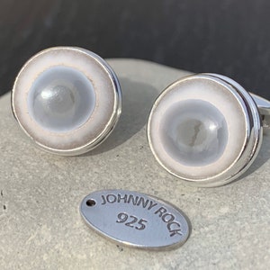 May include: A pair of silver cufflinks with a white and grey circular design. The cufflinks have a silver band around the edge and a smaller silver circle in the centre. The text "JOHNNY ROCK 925" is visible on a silver tag.