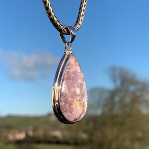 K&ouml;nnte beinhalten: Eine silberne Tropfen-Anh&auml;ngerkette mit einem rosafarbenen und wei&szlig;en Edelstein. Der Anh&auml;nger h&auml;ngt vor einem blauen Himmel.