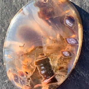 May include: A polished oval agate cabochon with brown, white, and pink patterns. The agate has a translucent appearance, revealing intricate layers of color.