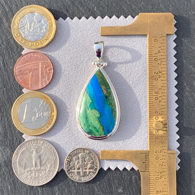 Puede incluir: Un colgante en forma de l&aacute;grima con una piedra preciosa azul y verde engastada en plata de ley. El colgante mide aproximadamente 2,5 cm de largo y se muestra junto a una regla y monedas para comparar el tama&ntilde;o.