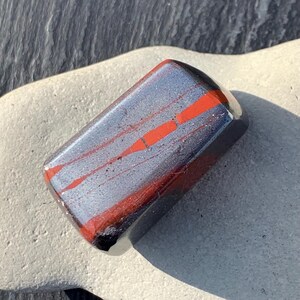 May include: A rectangular polished stone with a metallic grey base and striking red streaks. The stone is smooth and reflective, with dark edges. It rests on a light grey surface. The stone's dimensions are approximately 5 cm in length.