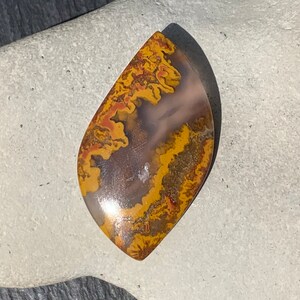May include: A polished, teardrop-shaped agate cabochon with a brown, yellow, and red pattern. The agate has a smooth, glossy finish.