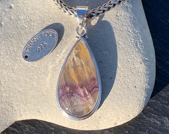 May include: A teardrop-shaped pendant with a silver setting and a chain. The pendant features a polished stone with shades of yellow, purple, and clear. A small silver tag with the text "JOHNNY ROCK 925" is also present.