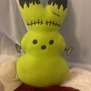 Può includere: Un peluche a forma di coniglietto Frankenstein verde lime. Il coniglietto ha capelli cuciti neri, occhi neri e dettagli cuciti neri. Ha due chiavi a carica argentate attaccate ai lati. Il peluche è su una superficie rossa e bianca.