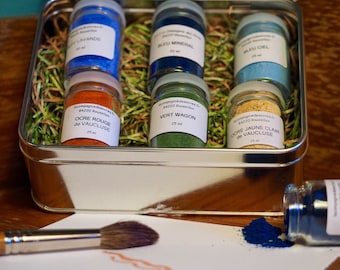 May include: A set of six small glass jars filled with different colored pigments. The jars are arranged in a metal tin with green grass-like material. The jars are labeled with French text and the names of the colors, including "Ocre Rouge de Vaucluse", "Vert Wagon", and "Ocre Jaune Clair de Vaucluse".