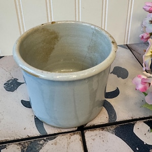 May include: A small, round, light blue ceramic pot with a slightly flared rim. The pot has a crack on the side and a worn, aged appearance. The interior is a lighter shade of blue. The pot is sitting on a patterned surface.