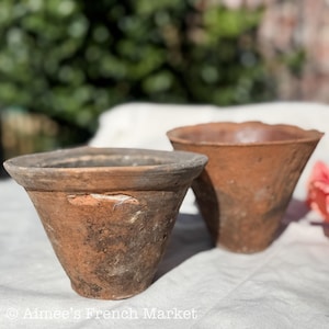 May include: Two weathered, terracotta-colored flower pots. The pots have a conical shape with a wide rim. One pot has a visible chip. The background is blurred, with greenery and a white cloth visible.