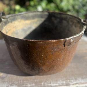 May include: A rustic, antique copper bucket with a rounded body and a slightly flared rim. The bucket has a dark, aged patina with visible wear and tear, and features small metal handles on either side for carrying. The interior shows signs of use.