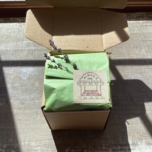 May include: A brown cardboard box with a green tissue paper lining. The box is open and a sprig of lavender is visible. A sticker with the text "Aimee's French Market" is on the box.