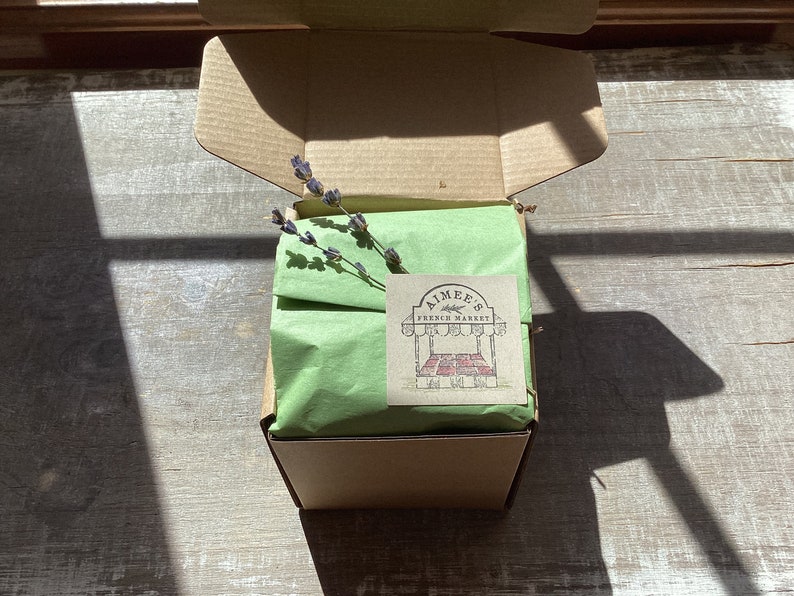 May include: A brown cardboard box with a green tissue paper lining. The box is open and a sprig of lavender is visible. A sticker with the text "Aimee's French Market" is on the box.
