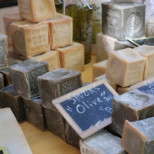 May include: A variety of rectangular bars of soap stacked on a wooden table. The soap is labeled "Savon de Marseille" and "Savons Olive 300g".