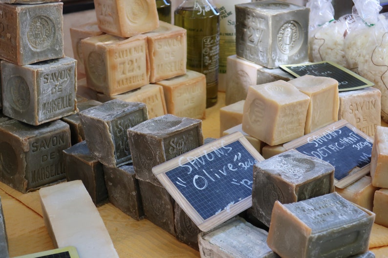 May include: A variety of rectangular bars of soap stacked on a wooden table. The soap is labeled "Savon de Marseille" and "Savons Olive 300g".