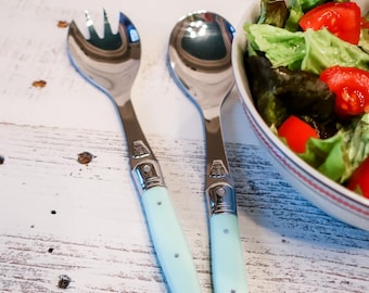 Könnte beinhalten: Ein Salatbesteck aus Edelstahl mit hellblauen Griffen. Jedes Besteckteil zeigt ein metallisches Bienenemblem am Kropf. Die Gabel hat drei Zinken und der Löffel eine abgerundete Laffe. Das Set liegt neben einer Schüssel mit Salat.