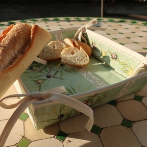 May include: A green and white fabric bread basket with a white fabric handle. The basket is filled with a baguette and sliced bread. The basket is sitting on a tiled surface.