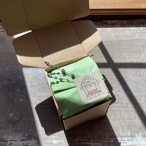May include: A brown cardboard box with a green tissue paper liner and a small sprig of lavender. The box is closed and has a sticker on the top that says "Aimee's Fresh Market".