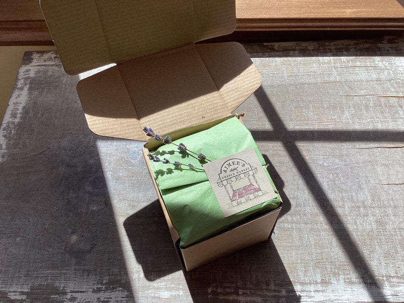 May include: A brown cardboard box with a green tissue paper liner and a small sprig of lavender. The box is closed and has a sticker on the top that says "Aimee's Fresh Market".