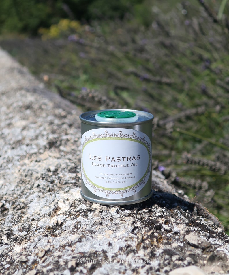 May include: A silver can of Les Pastras Black Truffle Oil, with a green lid. The can is sitting on a gray, textured surface. The label reads "Les Pastras Black Truffle Oil", "Tuber Melanosporum", "Organic Product of France", and "9 ml / 3 fl oz".