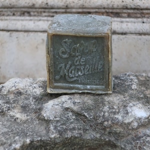 May include: A bar of green soap with the text "Savon de Marseille" on the front. The soap is sitting on a rough, gray stone surface.