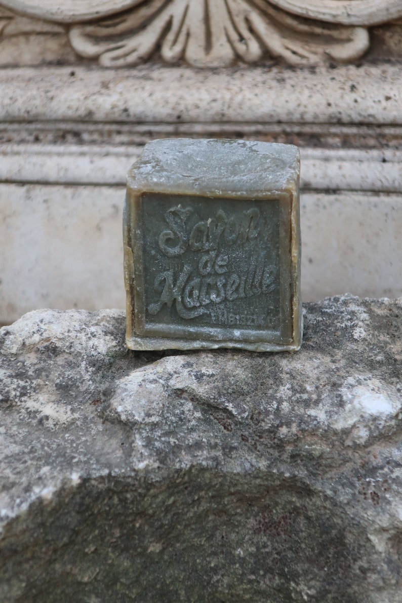 May include: A bar of green soap with the text "Savon de Marseille" on the front. The soap is sitting on a rough, gray stone surface.