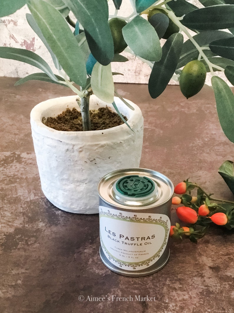 May include: A small silver can of Les Pastras Black Truffle Oil with a green lid. The can is sitting on a brown surface next to a potted olive tree with green olives.
