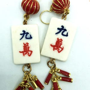 May include: Gold-tone earrings featuring red and gold accents. Each earring has a red lantern-shaped bead, a white tile with Chinese characters, and a cluster of red and gold charms. A unique accessory.