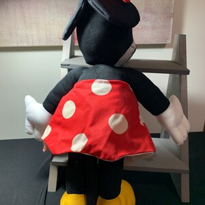 May include: A plush Minnie Mouse doll, viewed from the back, features a black body, white gloves, and yellow shoes. The doll wears a red skirt with white polka dots and a matching red bow with white polka dots. The doll is positioned in front of a gray wooden shelf.