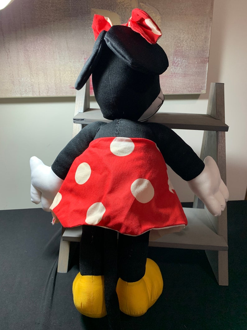 May include: A plush Minnie Mouse doll, viewed from the back, features a black body, white gloves, and yellow shoes. The doll wears a red skirt with white polka dots and a matching red bow with white polka dots. The doll is positioned in front of a gray wooden shelf.