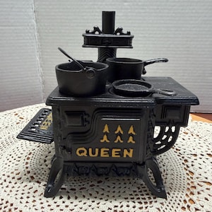 May include: A miniature black cast iron toy stove with gold-painted accents. The front features the word QUEEN and decorative patterns. It includes a small pot, a saucepan, and a skillet on the stovetop, with a side shelf and a chimney stack.