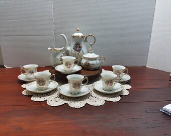 May include: A vintage porcelain tea set with a floral design. The set includes a teapot, sugar bowl, creamer, and six teacups with saucers. Gold trim accents the white and pastel-colored pieces. The set is arranged on a lace doily.