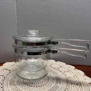 May include: A clear glass double boiler with a lid and two clear handles. The pot has a silver band around the top and bottom. The double boiler sits on a cream-colored, crocheted doily.