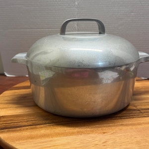 Puede incluir: Una olla redonda de metal color plateado con su tapa a juego. La olla cuenta con dos asas laterales y un asa arqueada en la parte superior de la tapa. La superficie exterior del metal muestra signos de desgaste y ligeros arañazos.