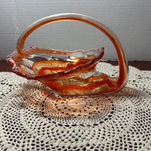 Puede incluir: Una cesta decorativa de vidrio con un cuerpo transparente y remolinos naranjas y rojos. La cesta tiene un asa y un borde festoneado. La cesta está sobre un tapete de ganchillo blanco.