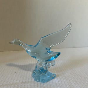 May include: A light blue glass figurine of a bird in flight. The bird has outstretched wings and is perched on a cloud-like base. The glass is transparent, allowing light to pass through.