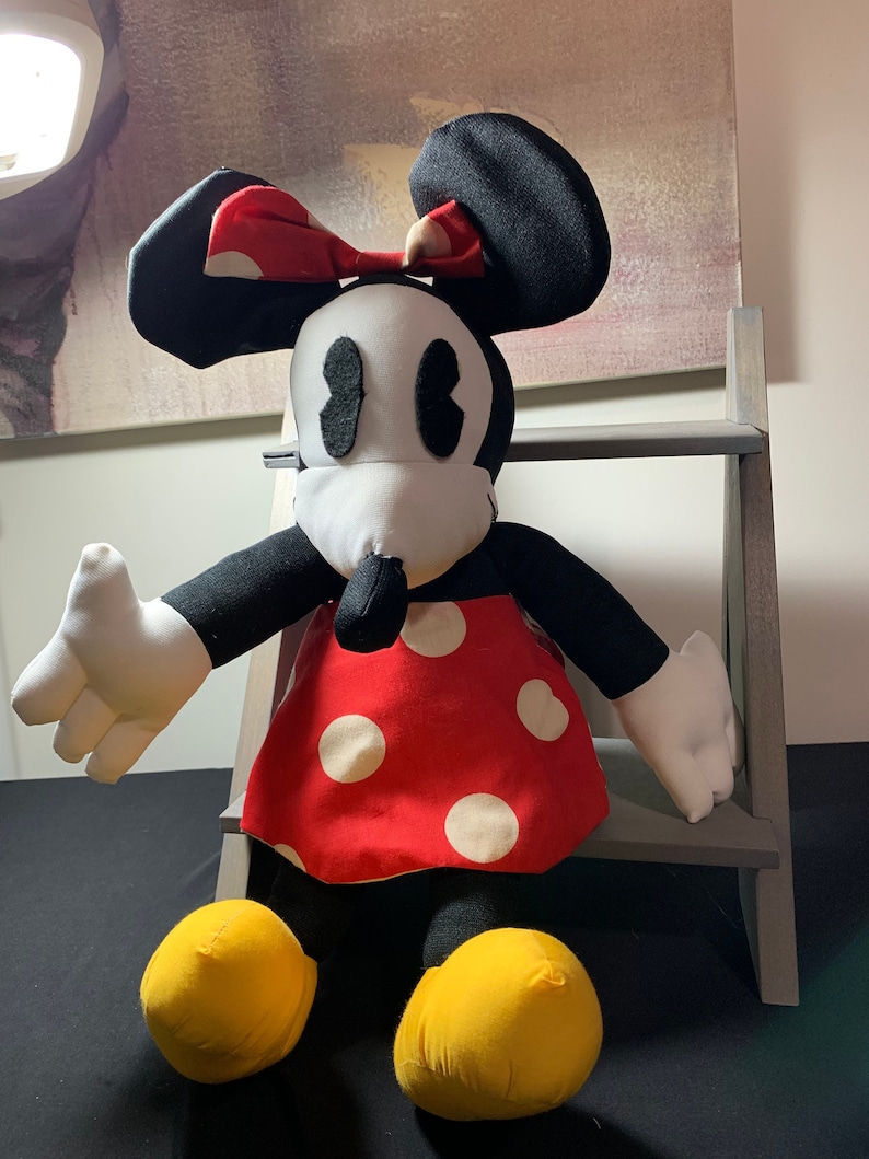 May include: A plush Minnie Mouse doll with a red dress with white polka dots, a red bow with white polka dots, and yellow shoes. The doll has black ears, arms, and legs, and a white face with black eyes and nose.