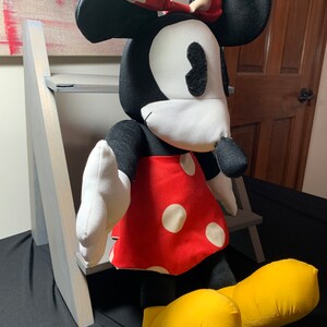 May include: A plush Minnie Mouse doll with a black head and ears, white face, and a red dress with white polka dots. The doll has a red bow with white polka dots and yellow shoes. The doll is sitting in front of a gray wooden structure.