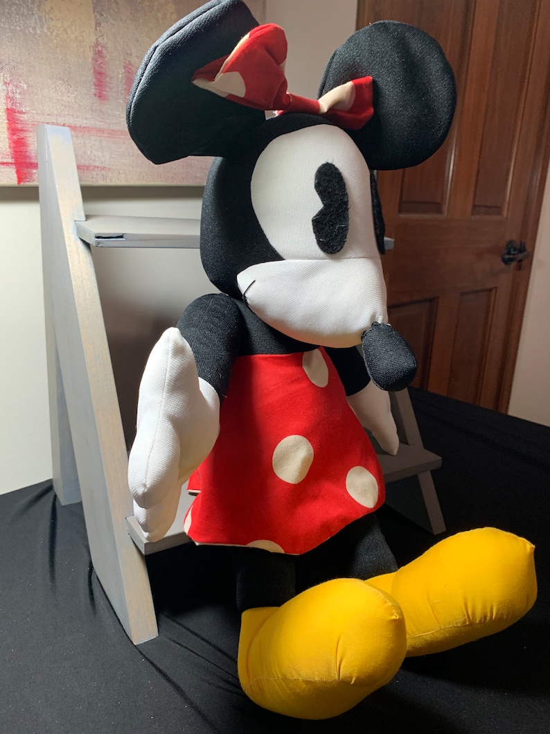May include: A plush Minnie Mouse doll with a black head and ears, white face, and a red dress with white polka dots. The doll has a red bow with white polka dots and yellow shoes. The doll is sitting in front of a gray wooden structure.