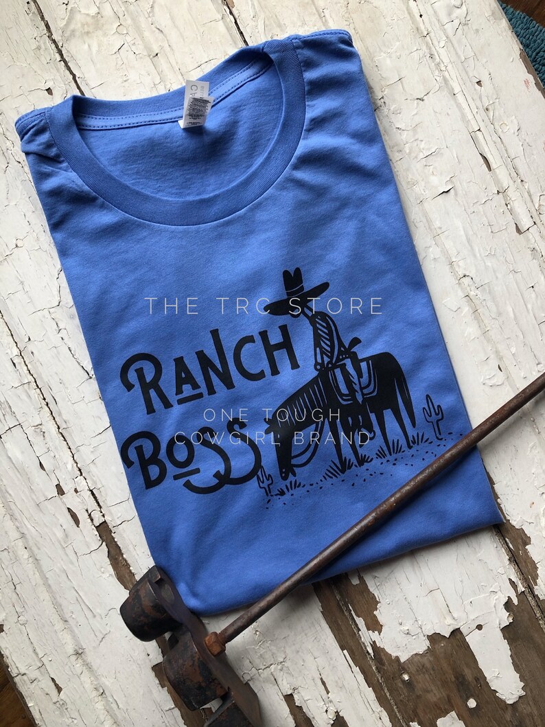 May include: A blue t-shirt with a black graphic design of a cowboy riding a horse. The graphic includes the text "The T-Roc Store Ranch Boss One Tough Cowgirl Brand".