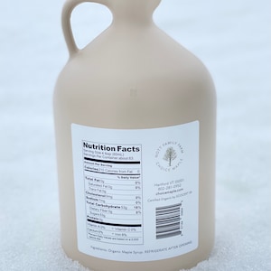 May include: A brown plastic jug of maple syrup with a black lid and a white label with the words "Nutrition Facts" and "Nott Family Farm Choice Maple" printed on it. The label also includes the company's address, phone number, and website. The jug is sitting on a white surface.