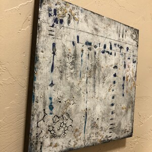 K&ouml;nnte beinhalten: Abstrakte Malerei auf Leinwand mit wei&szlig;em Hintergrund und blauen und goldenen Akzenten. Das Gem&auml;lde hat eine strukturierte Oberfl&auml;che und einen verwitterten Look.