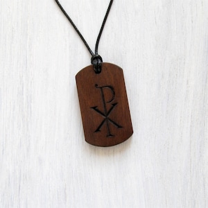 Peut inclure: Un collier pendentif en bois brun foncé avec un cordon noir. Le pendentif est rectangulaire avec des bords arrondis et comporte un symbole Chi Rho sculpté. Le collier est présenté sur un fond en bois blanc.