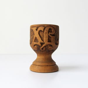 May include: A carved wooden egg cup with a pedestal base. The cup is a light brown colour and features the carved letters "XA" on the side. The design is detailed and the cup has a smooth finish.