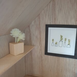 May include: A white wooden shelf with a small white vase with white flowers on it. A framed black and white print of potted plants hangs on the wall behind the shelf.