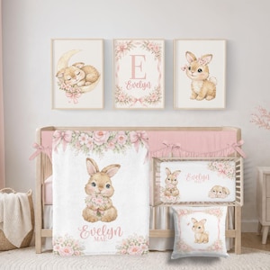 Könnte beinhalten: Ein Kinderbett-Set bestehend aus einer Decke, einem Bettlaken und einem Kissen mit Aquarellillustrationen von Hasen, rosa Blumen und dem Namen Evelyn. Drei gerahmte Wandbilder zeigen einen schlafenden Hasen, den Buchstaben E und einen sitzenden Hasen.