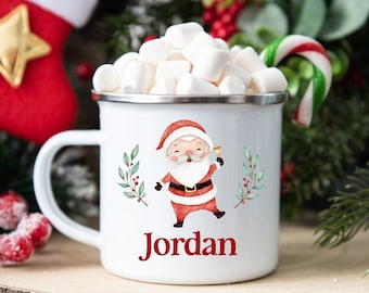Könnte beinhalten: Weiße Email Tasse mit einer roten und weißen Weihnachtsmann-Illustration, grünen Stechpalmenzweigen und dem Namen "Jordan" in rot gedruckt. Die Tasse ist mit weißen Marshmallows und einem roten und weißen Zuckerstangen gefüllt.
