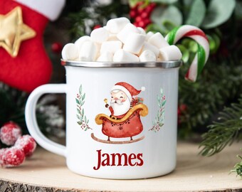 Könnte beinhalten: Weißer Emaillebecher mit Marshmallows, mit einem Weihnachtsmann-Design in einem Schlitten und dem Namen "James" in Rot. Der Becher hat einen silbernen Rand und einen weißen Griff. Festliches Weihnachtsthema.