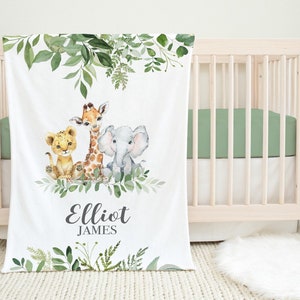 Peut inclure: Couverture de bébé blanche avec une illustration à l'aquarelle d'un lionceau, d'une girafe et d'un éléphant entourés de verdure. La couverture est personnalisée avec le nom "Elliot James". La couverture est posée sur un lit bébé en bois clair.
