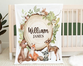 May include: White baby blanket with a woodland theme. The blanket features a brown tree stump with the names "William James" written on it.  The tree stump is surrounded by green leaves, a brown bear, a fox, a raccoon, a deer, and a squirrel.
