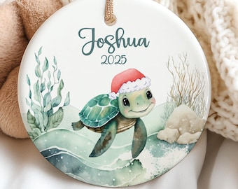May include: A beige teddy bear is positioned beside a round ceramic ornament. The ornament displays a watercolour illustration of a turtle wearing a Santa hat, with the name "Joshua" and the year "2025" above the turtle.