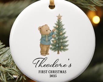 May include: A white ceramic Christmas ornament with a watercolor illustration of a teddy bear decorating a Christmas tree. The text "Theodore's FIRST CHRISTMAS 2025" is written in elegant script. The ornament hangs from a gold string.
