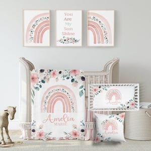May include: A nursery set with a cot, bedding, and wall art. The set includes a cot, a blanket, a pillow, and framed prints. The designs feature watercolour rainbows and floral accents in shades of pink and peach.